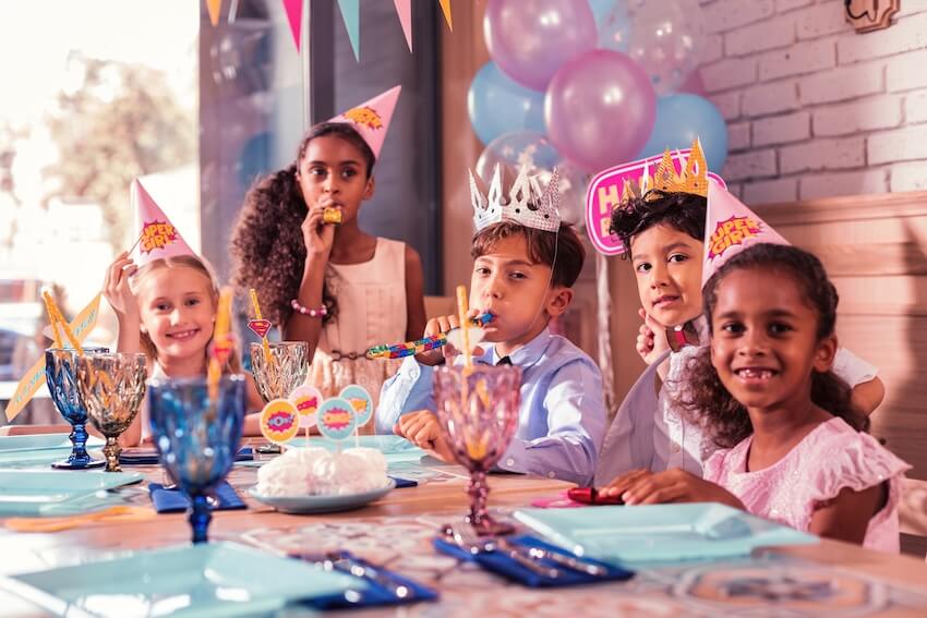 Birthday wishes for kids: 5 children wearing party hats and blowing party horns while smiling at the camera