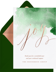 A holiday card featuring a handwritten script reading "joy" in a rose gold foil. Behind the word "joy" there is a splash of green watercolor.
