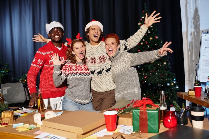Holiday party games: four friends wearing Christmas sweaters, smiling at a camera