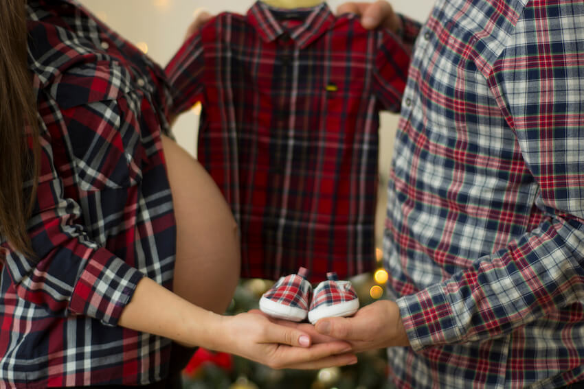 Christmas pregnancy announcement: husband and a pregnant woman holding their baby's clothes and shoes