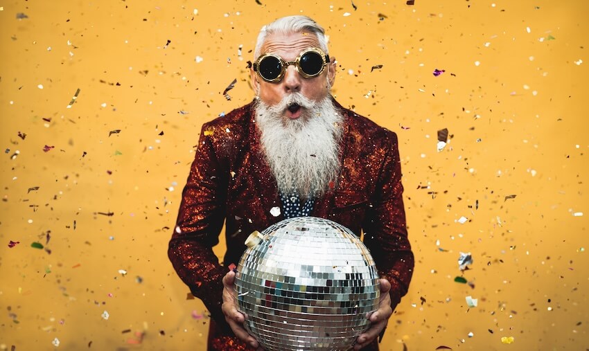 65th birthday ideas: senior man wearing a pair of sunglasses, holding a large disco ball, with confetti falling in the background